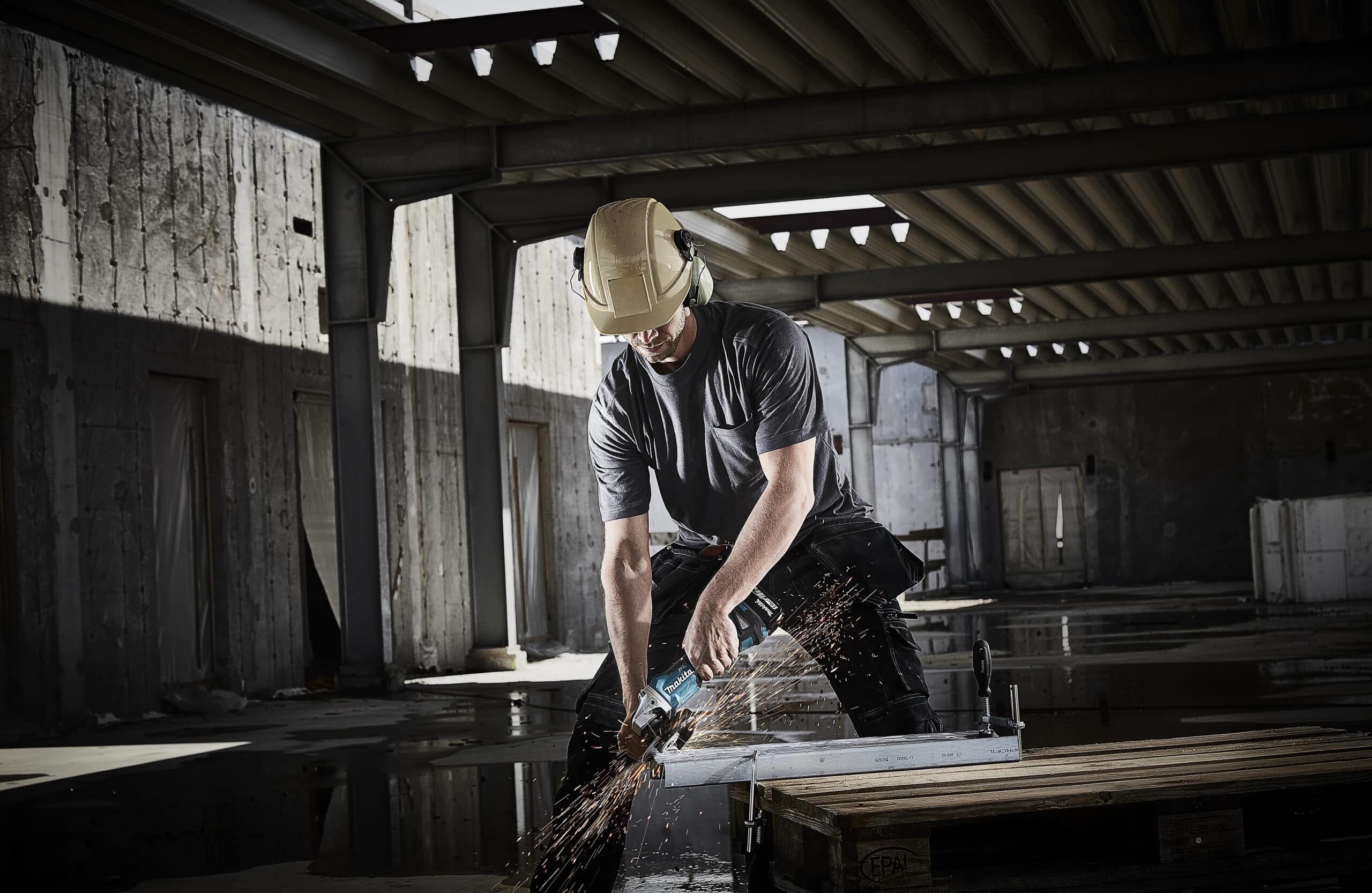 Construction worker wearing a yellow hard hat and safety glasses cuts a metal beam with a power grinder, sparks flying in a dim industrial space