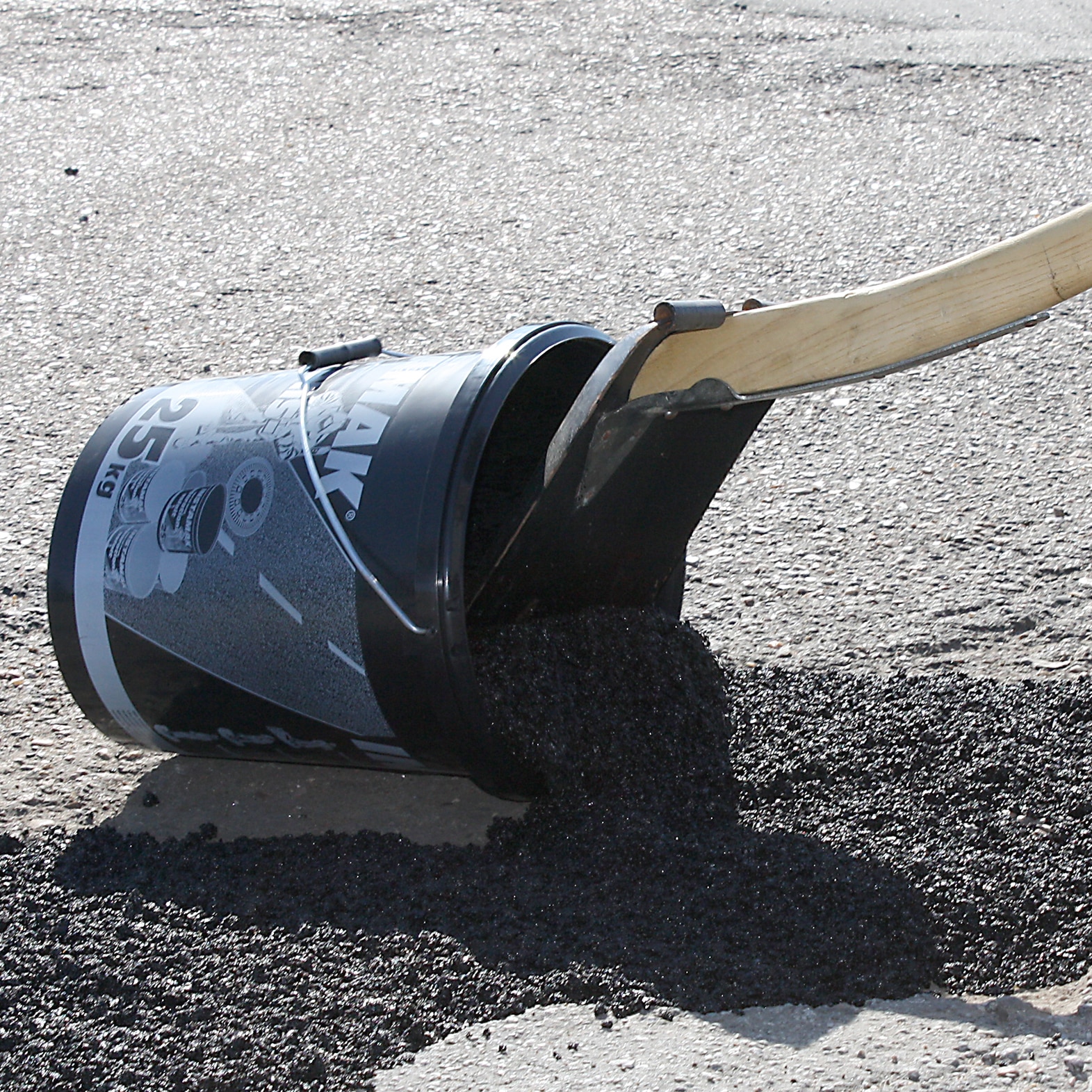 Overturned bucket of asphalt sealant spilling black tar onto a paved road, with a wooden-handled spreader nearby