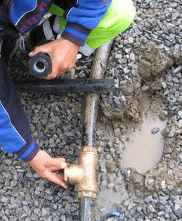 Person repairs an outdoor water pipe, turning a brass valve with a wrench while a second tool rests nearby on gravel.