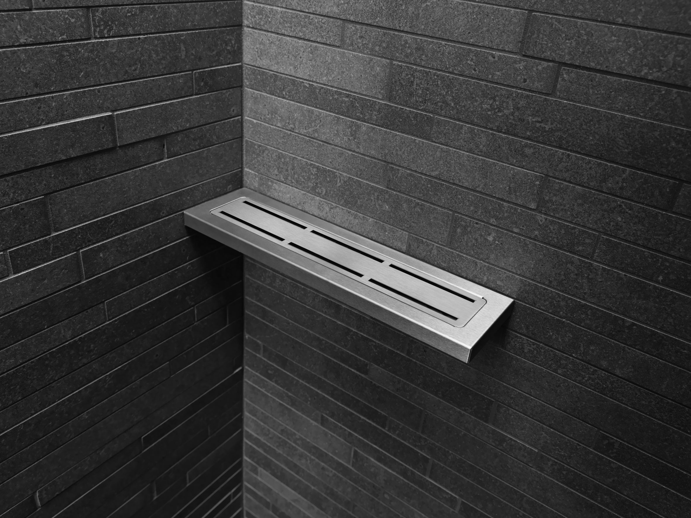Slim metal slatted bench mounted to a dark brick wall in a corner of a room.