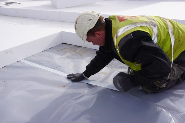 Construction worker in a hard hat and high-visibility vest kneels on a white roof, taping down a gray plastic moisture barrier.