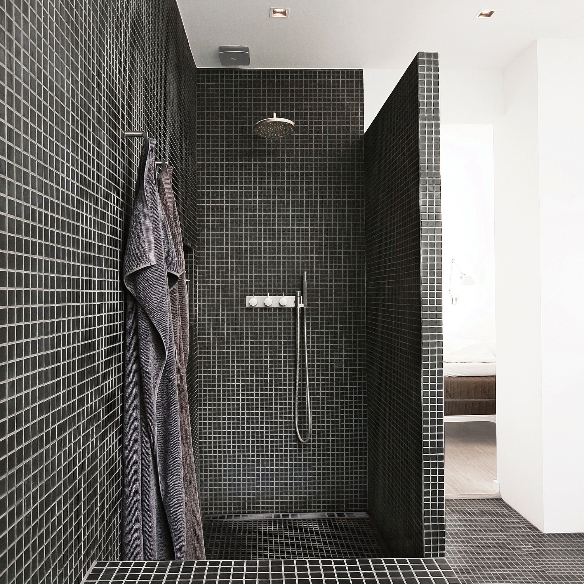 Modern shower area with black square-tiled walls, rainfall showerhead, and grey towels hanging on a rail.