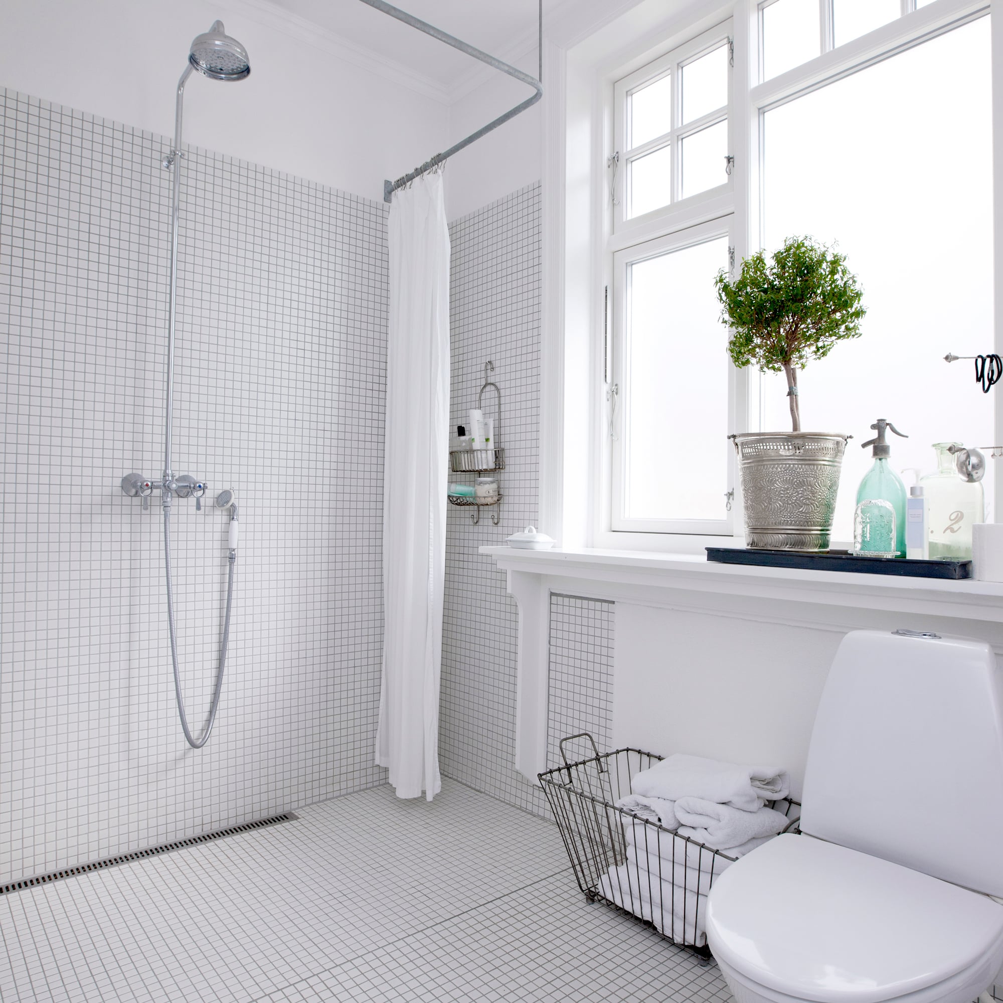 Bright bathroom with white grid-tile walls, a chrome showerhead and handheld shower, and a large window near a vanity with towels and a potted plant.