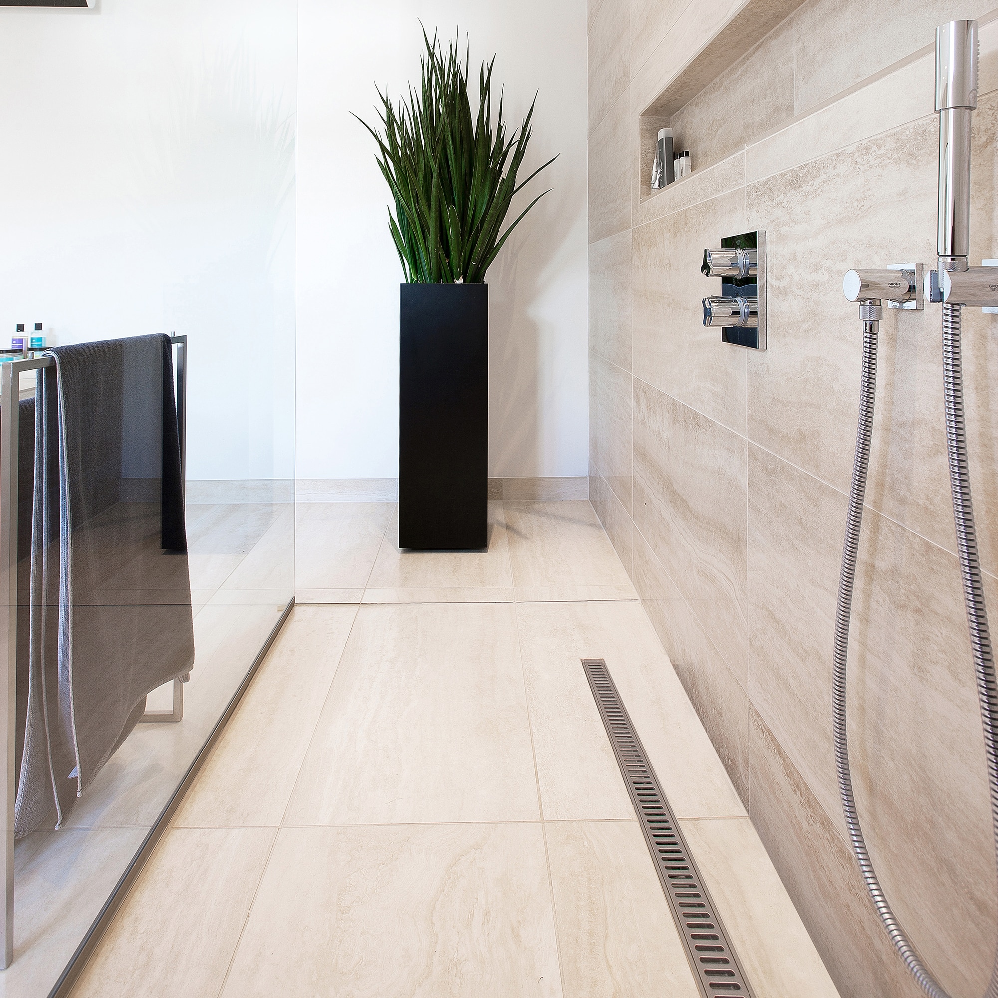 Modern bathroom featuring a tall black rectangular planter with a green plant, beige tiled walls, and a glass shower door on the left; built-in shower niche with toiletries and chrome fixtures visible on the wall.