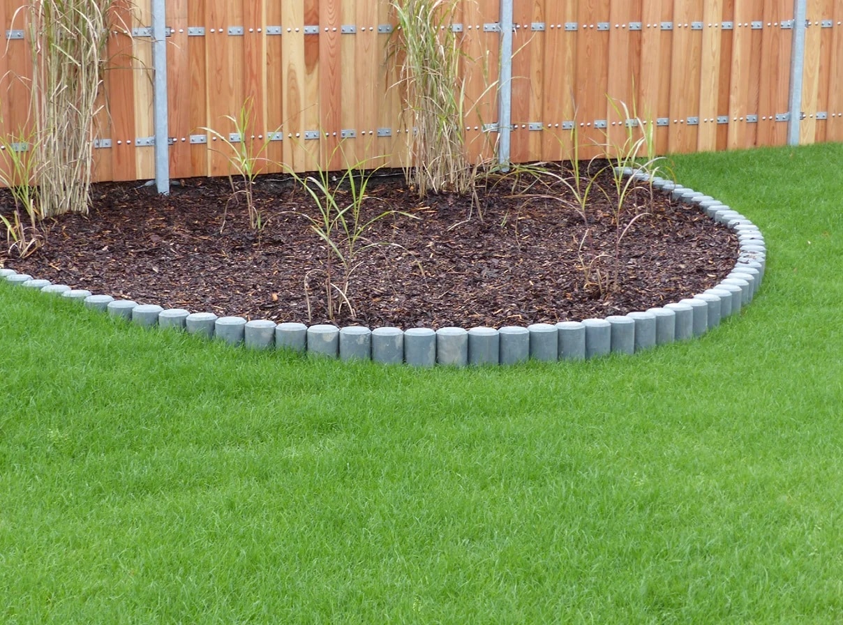 Curved garden bed with grey short concrete edging, mulch, and young grasses/plants beside a wooden fence and green lawn by a metal fence stake.