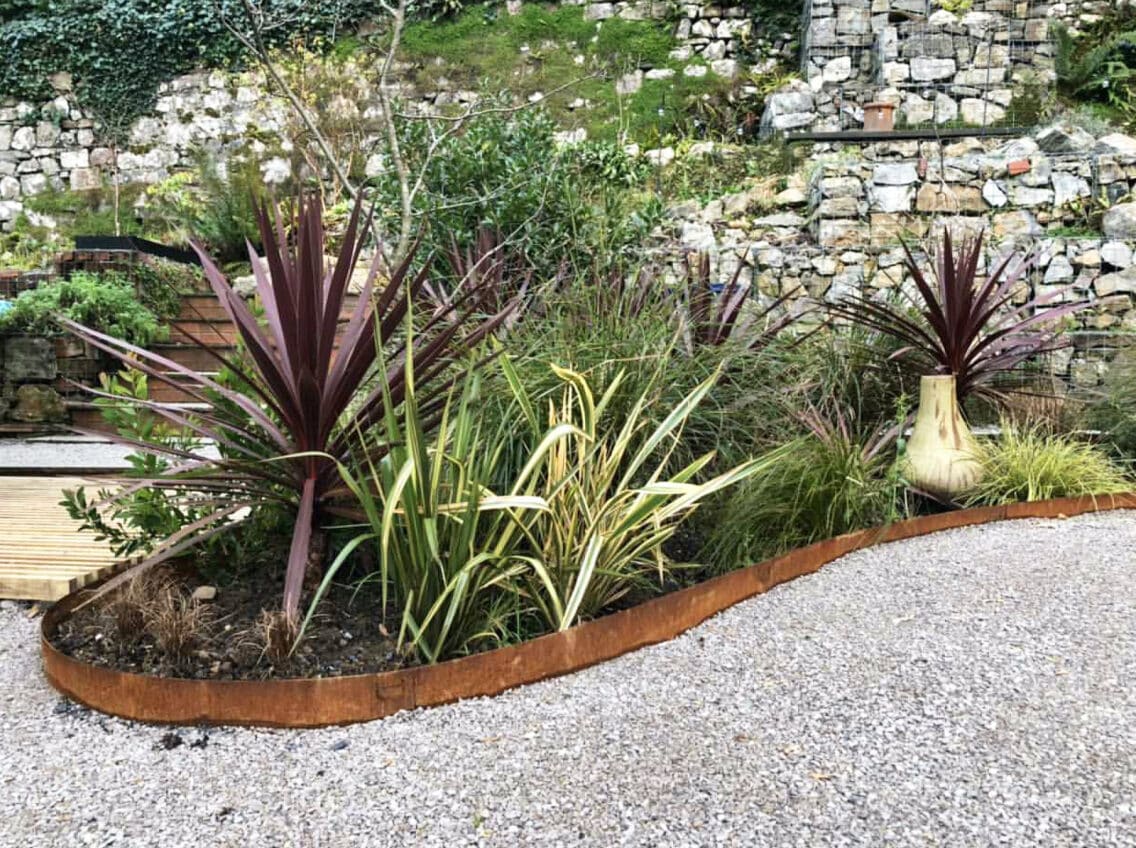 Curved rusty metal edging around a plant bed with purple-leafed and green grasses beside a gravel path.",