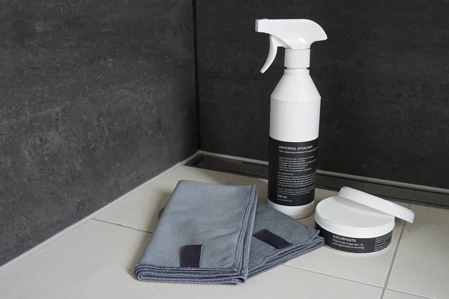 White spray bottle labeled 'UNIVERSAL AFKALKER', a jar labeled 'NATURPASTA', and folded blue microfiber cloths on a light tiled surface.