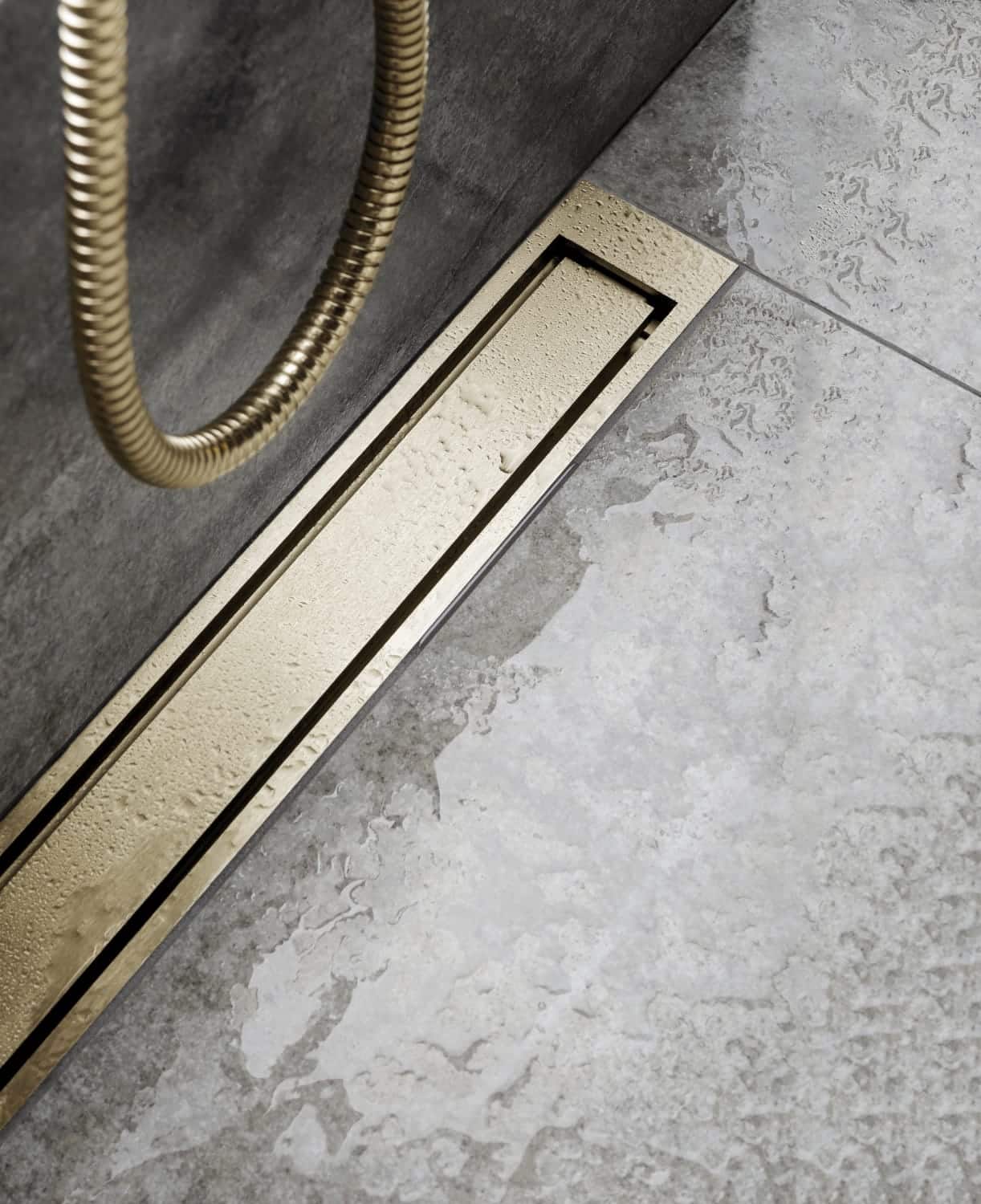 Gold rectangular floor drain set into a gray tiled bathroom floor, with a coiled flexible hose nearby and water droplets on the drain cover.