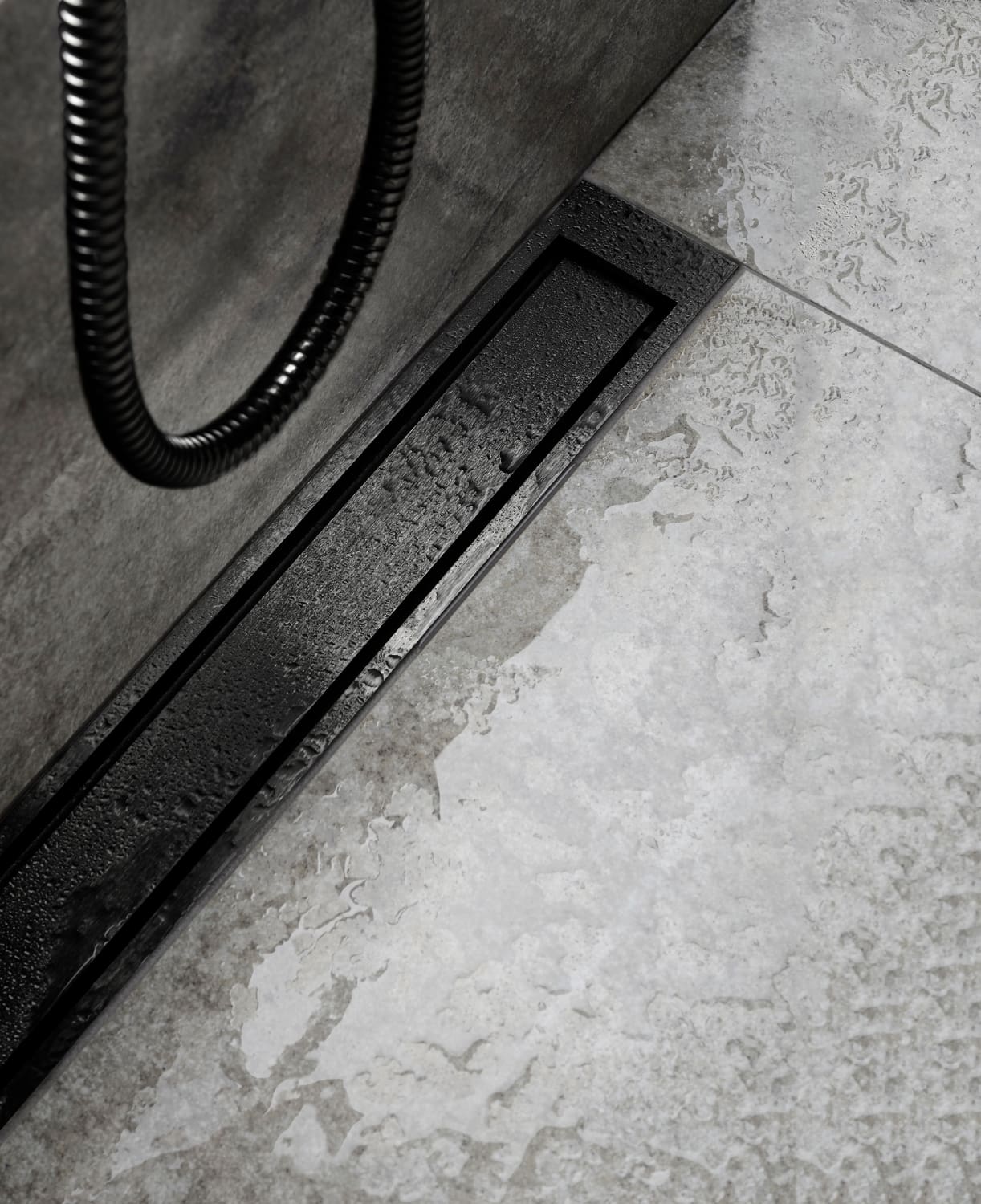 Close-up of a black rectangular shower drain set in a wet gray tile floor, with a coiled hose nearby.