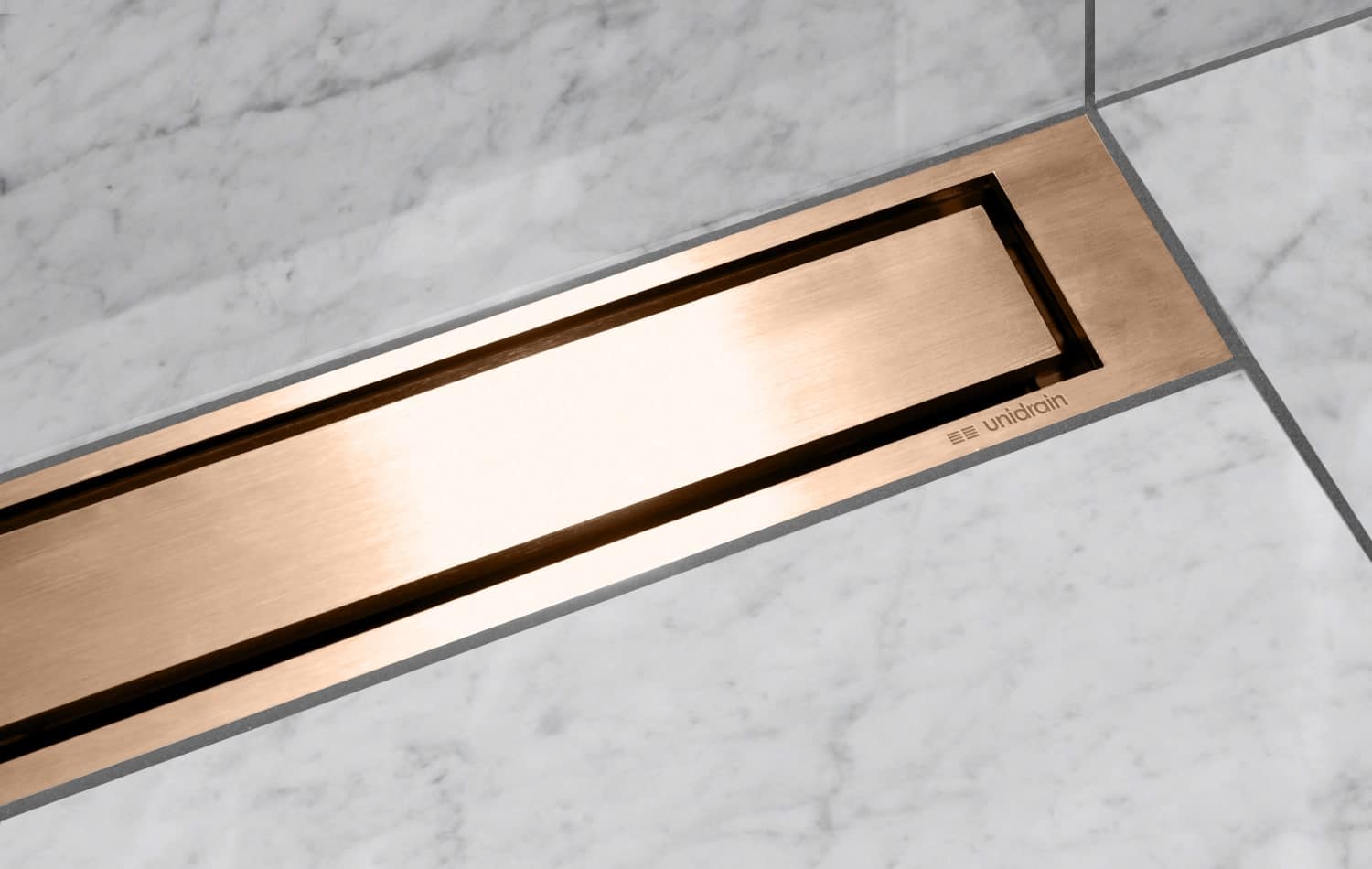 Copper-toned rectangular floor drain set in light gray marble tiles (Unidrain)