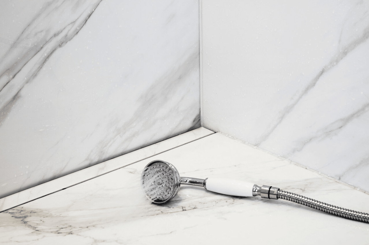 Handheld chrome showerhead with white handle and flexible hose resting on a marble shower ledge.