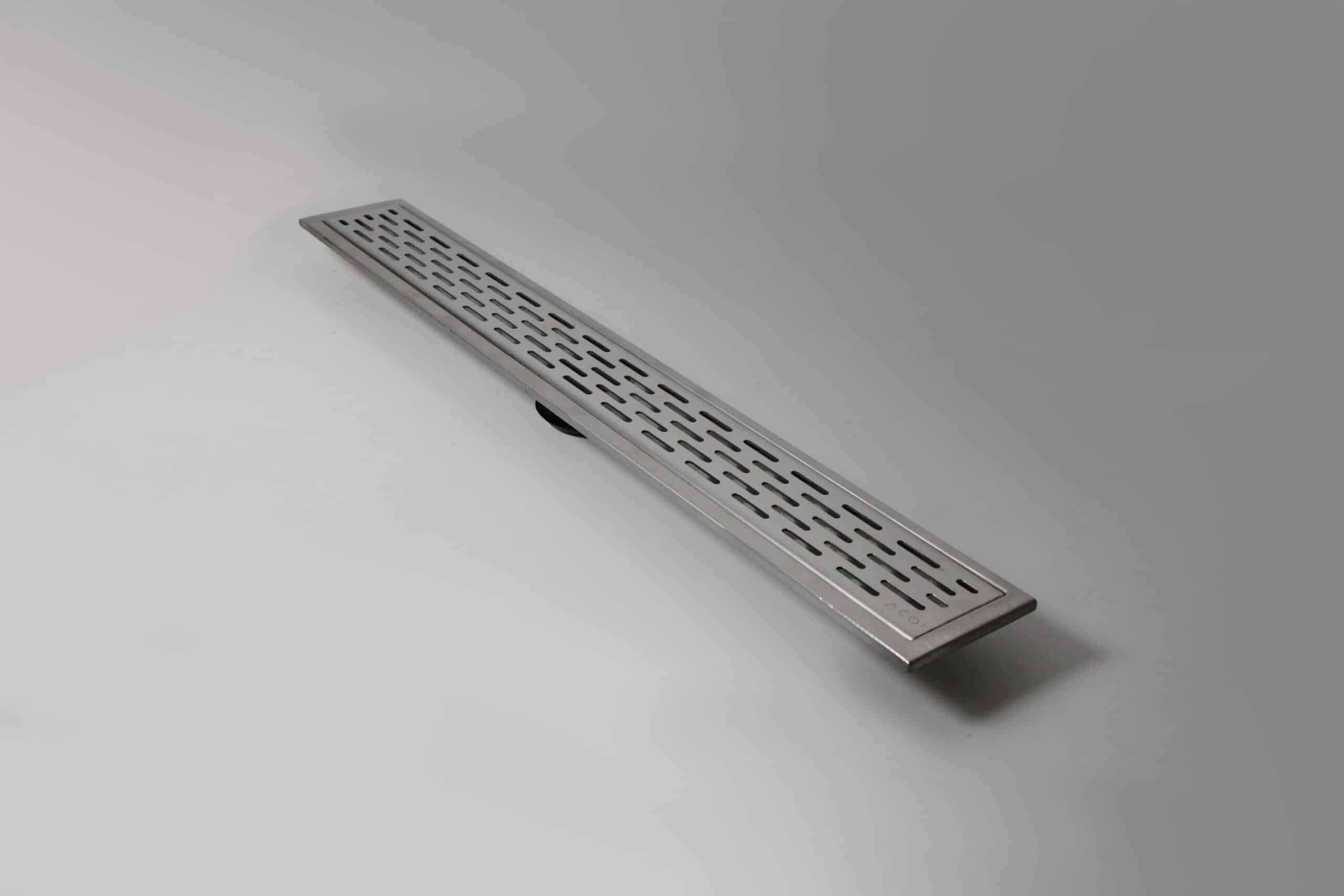 Stainless steel linear shower drain with perforated slots lying diagonally on a light surface, viewed from above