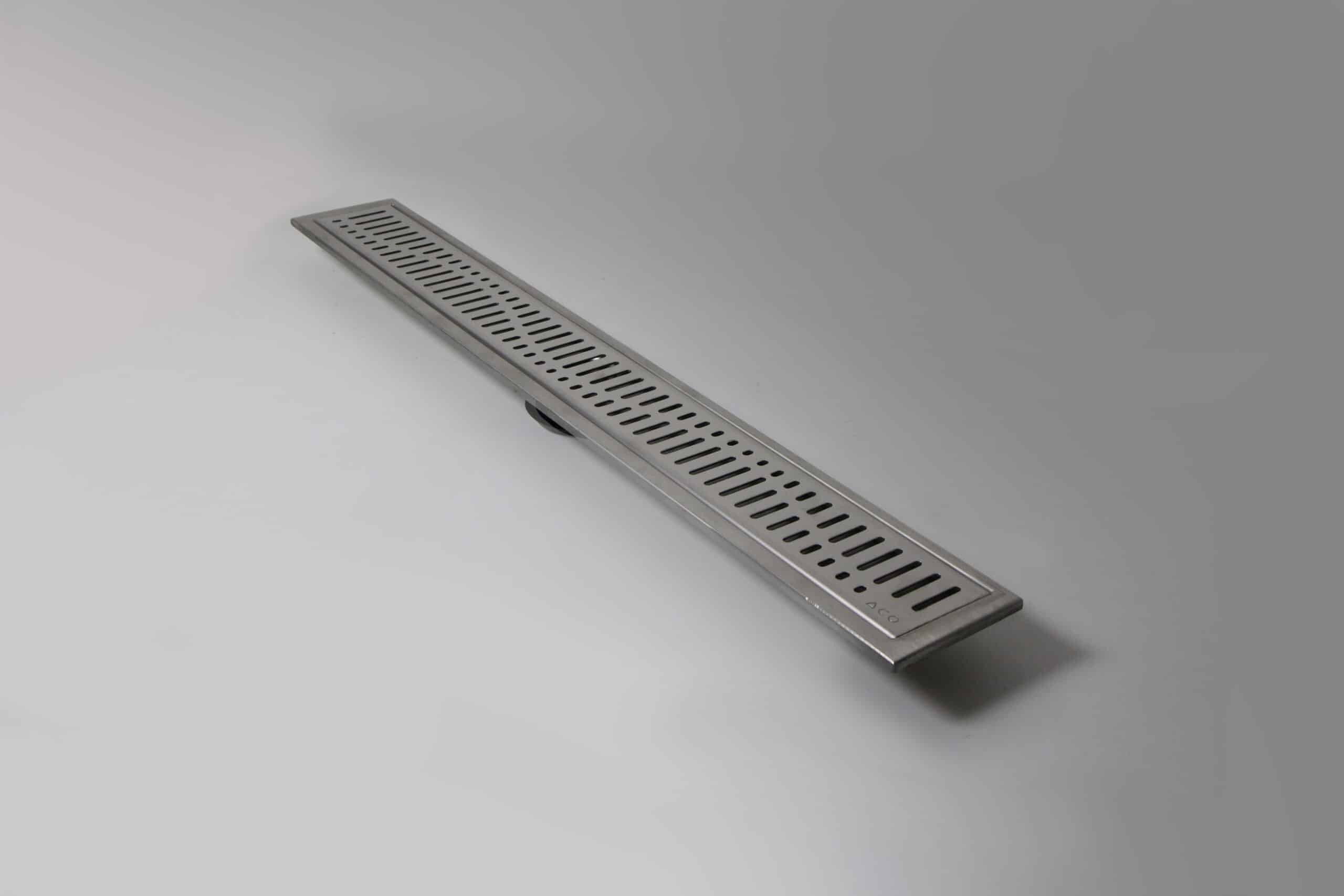 Elongated stainless steel floor drain with rectangular slots on a light gray surface