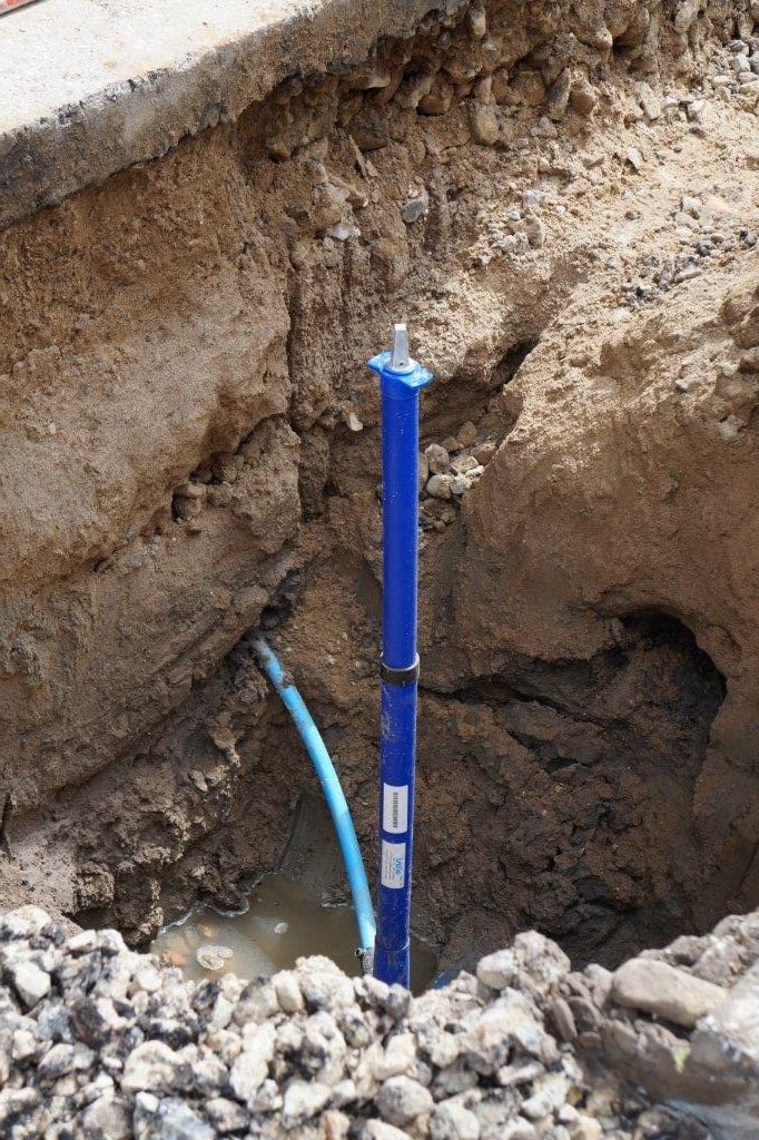 Blue PVC utility pipe stands upright in a deep trench with muddy soil and rubble, a light-blue hose nearby.