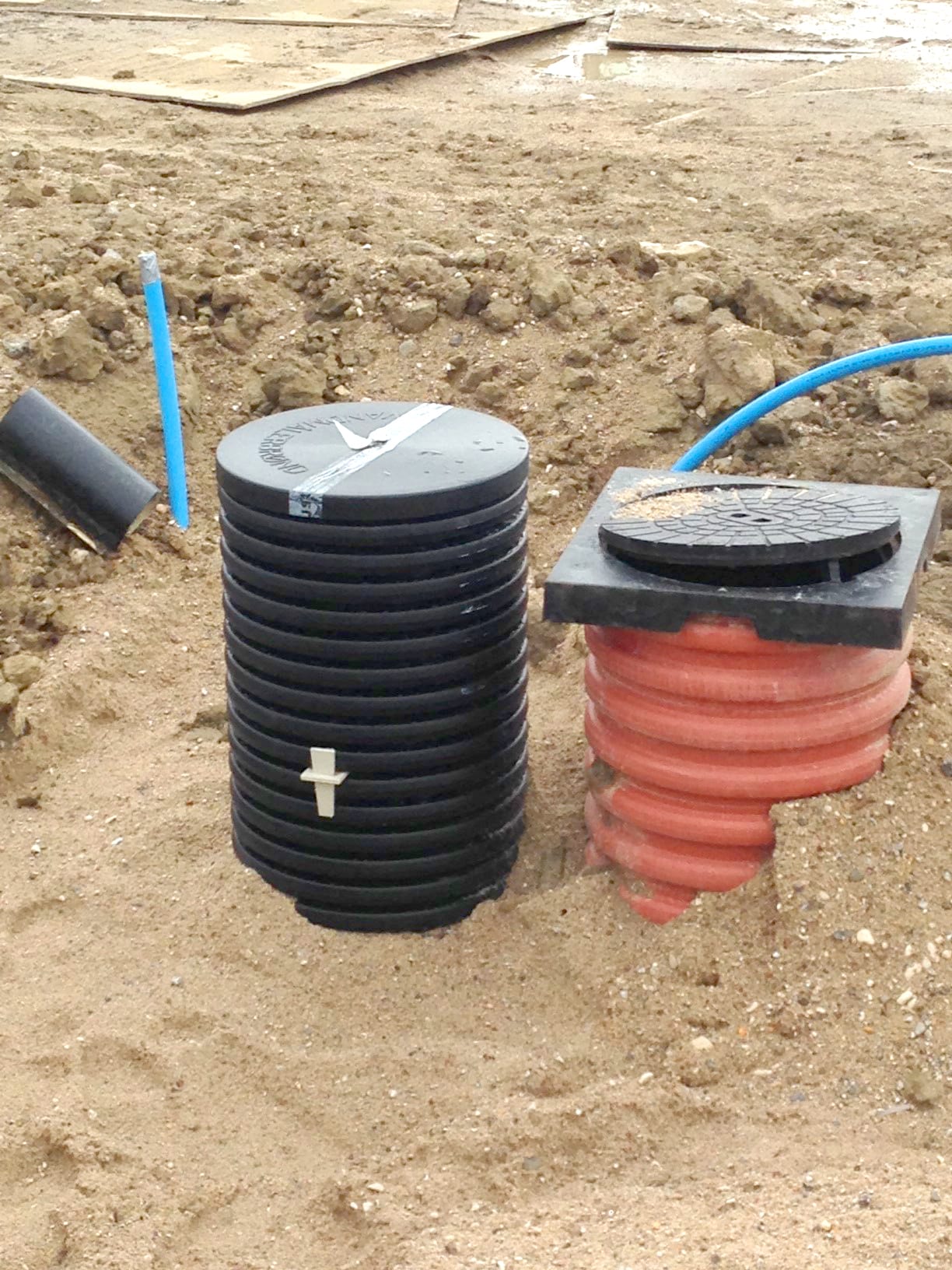 Construction site showing underground drainage components: stacked black corrugated pipes and a red corrugated pipe with a manhole lid, blue hose nearby.