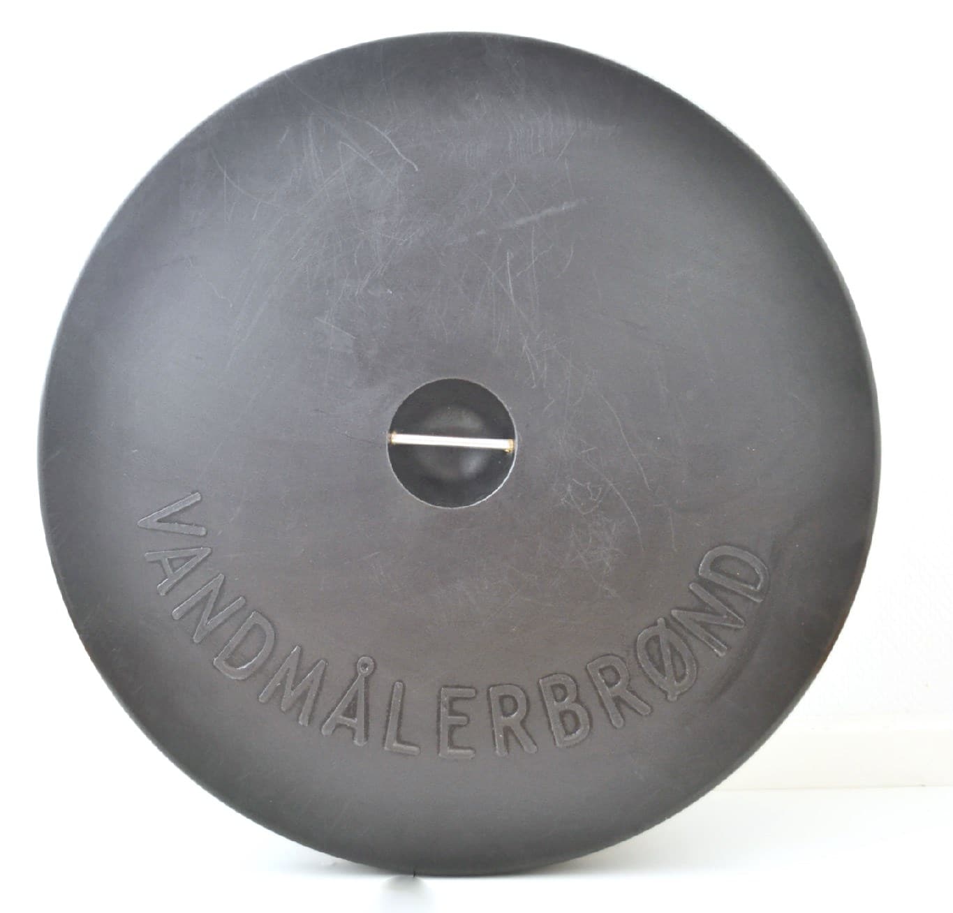Round black circular lid with a small central opening and a horizontal metal pin; embossed text around the edge reads 'VANDMÅLERBROEN'