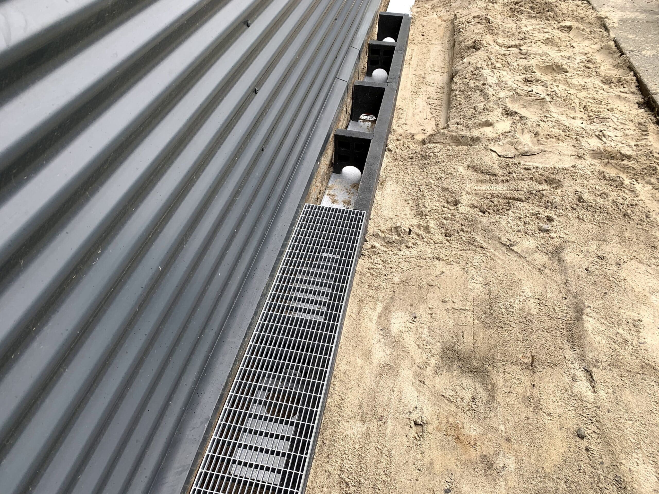Construction site scene: gray corrugated metal wall, row of black hollow blocks with white spheres, a metal grate, and sandy soil beside a trench.