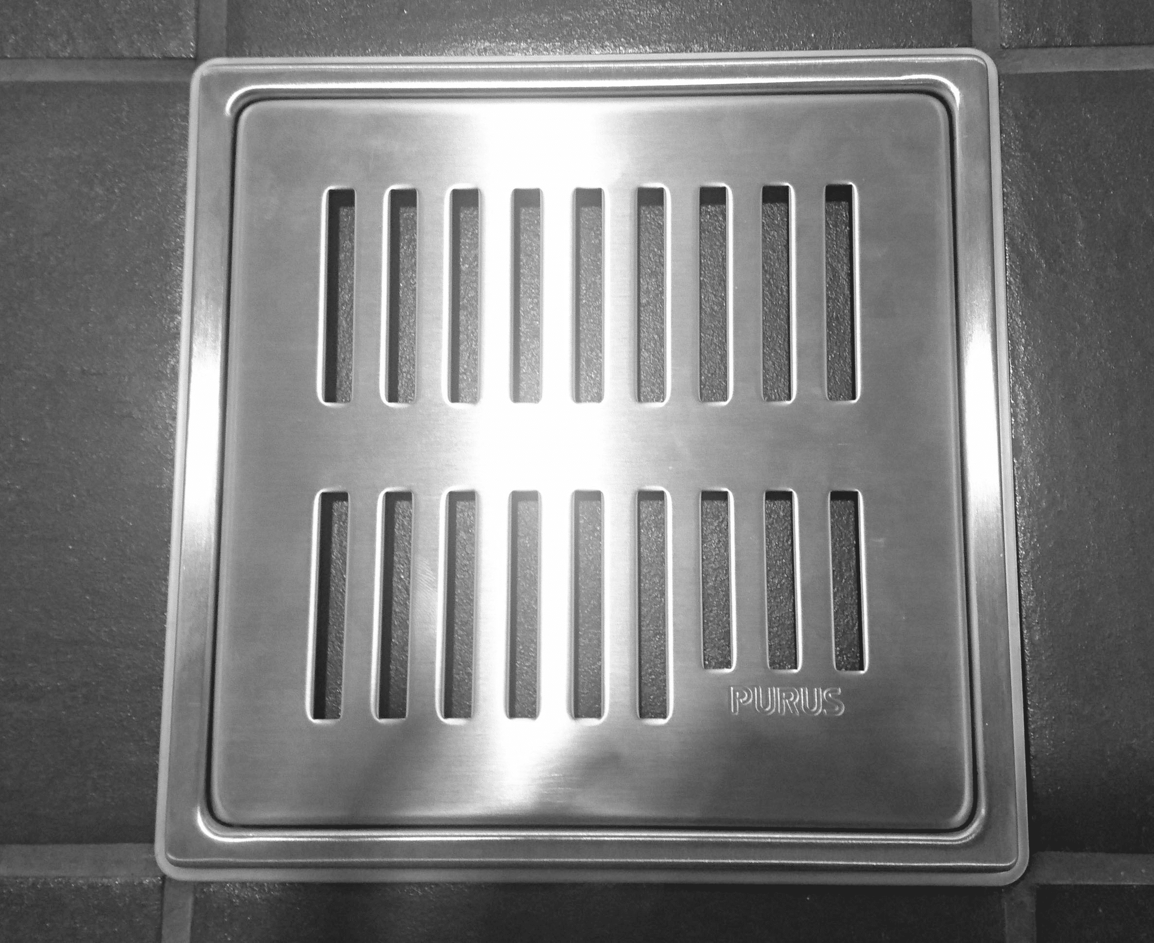 Metal floor drain cover with vertical rectangular slots on dark tiled floor.